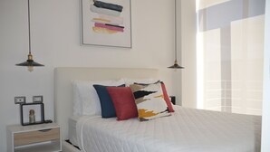 Una cama bien hecha con colcha blanca, una mesita de noche con lámpara y una obra de arte en la pared sobre la cama.