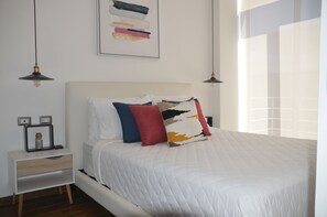 A neatly made bed with a white quilt, a bedside table with a lamp, and a wall art piece above the bed.