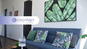 Una sala de estar moderna con un sofá azul, cojines estampados de motivos tropicales, un cuadro de hojas verdes y una jaula decorativa sobre una mesita.