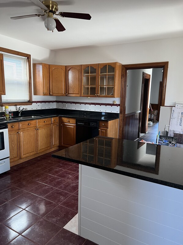 A kitchen with wooden cabinets, a black countertop, a ceiling fan, and a window with blinds. - Colonial sky light very large rooms (Boston)