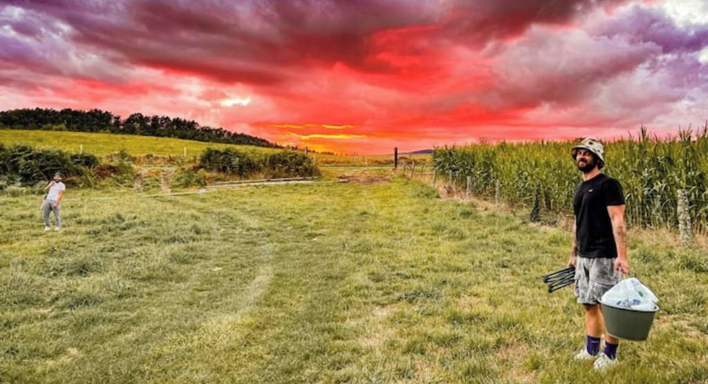 A field with a person holding a bucket and a tray of items, another person walking in the distance, and a dramatic sunset sky.