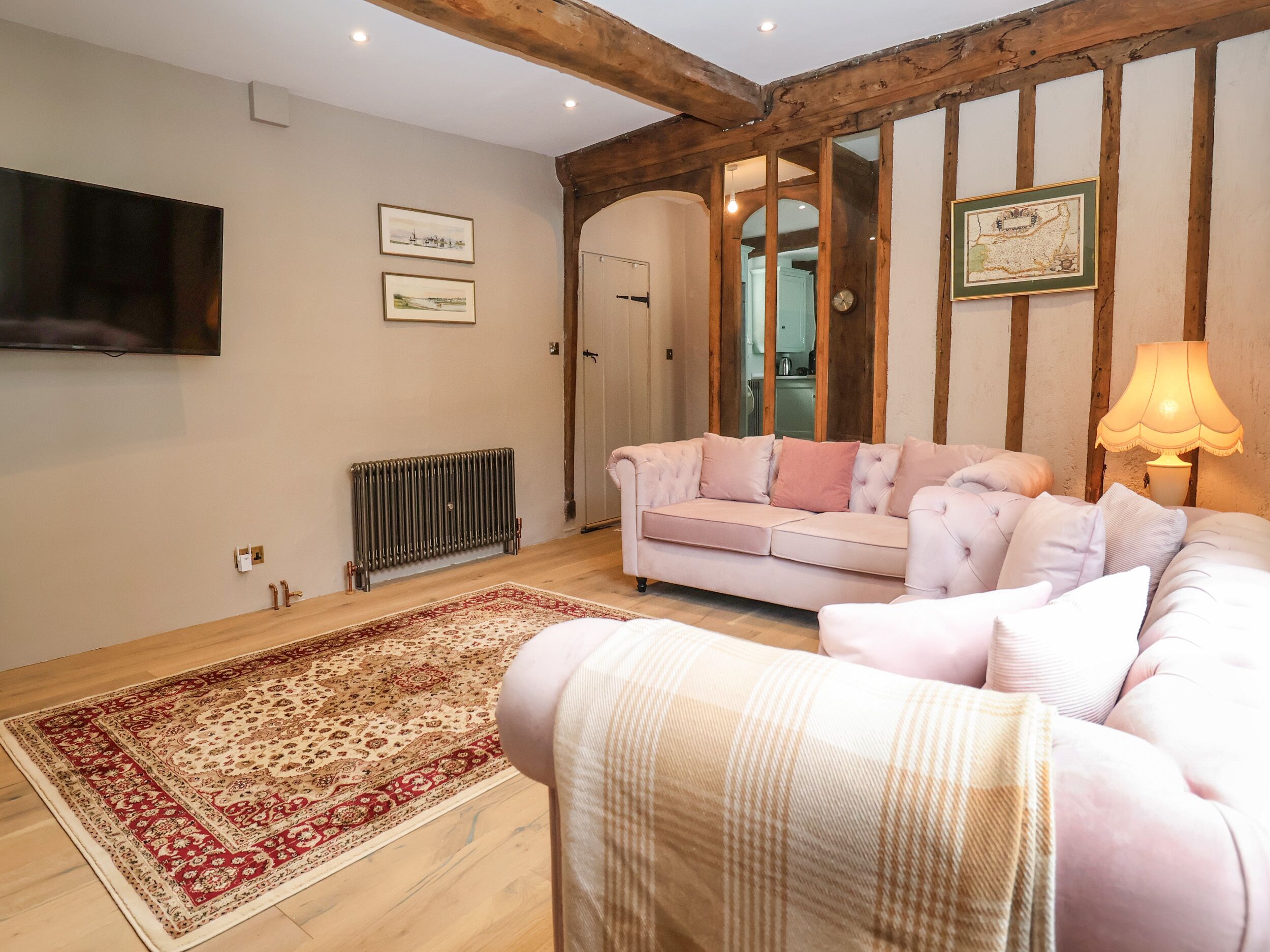 A cozy living room with a pink sofa, a patterned rug, wooden beams, a radiator, and a TV mounted on the wall.