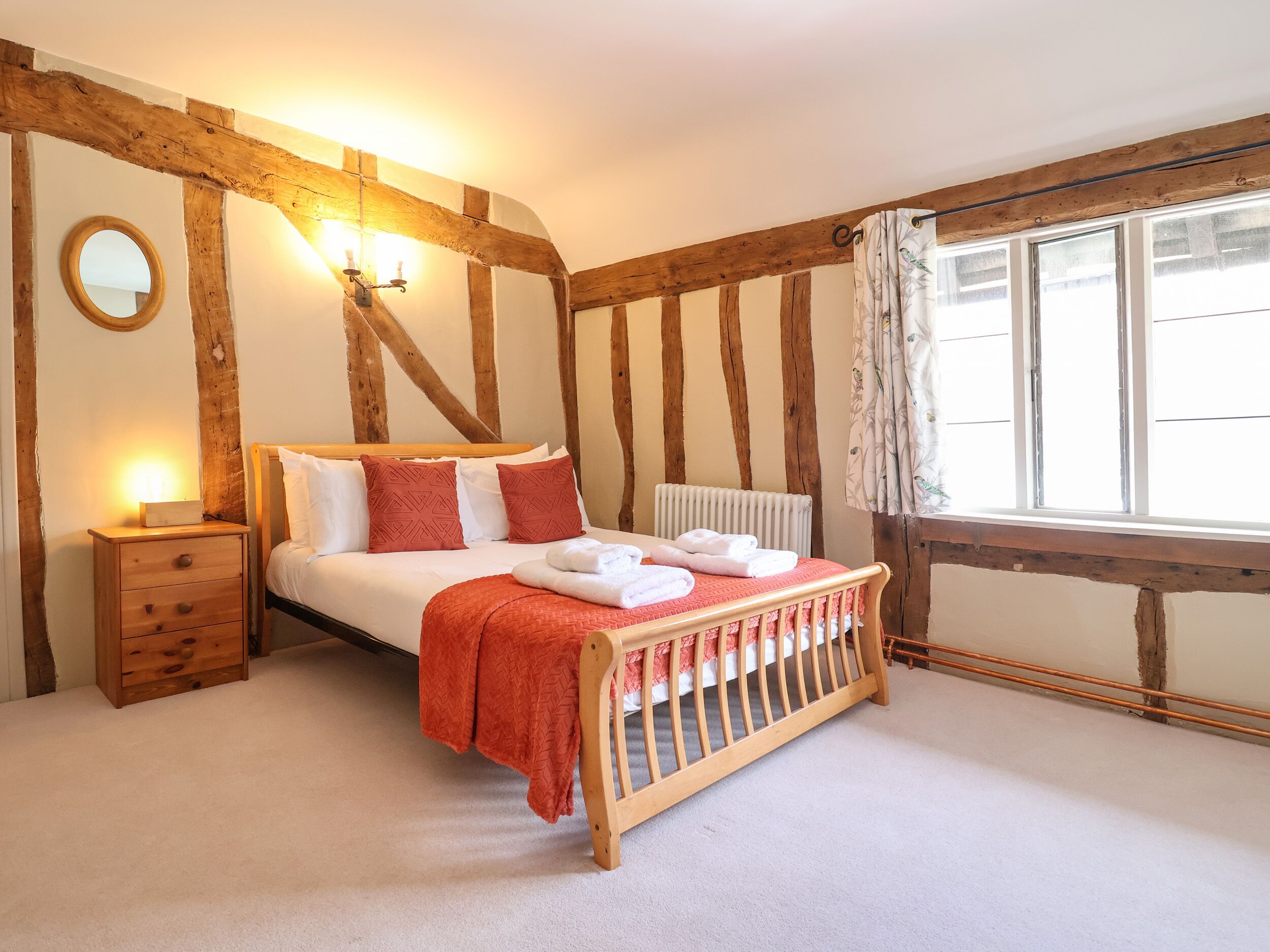 Un dormitorio con vigas de madera a la vista, una cama de madera con una manta roja, una mesita de noche con lámpara y una ventana con cortinas.