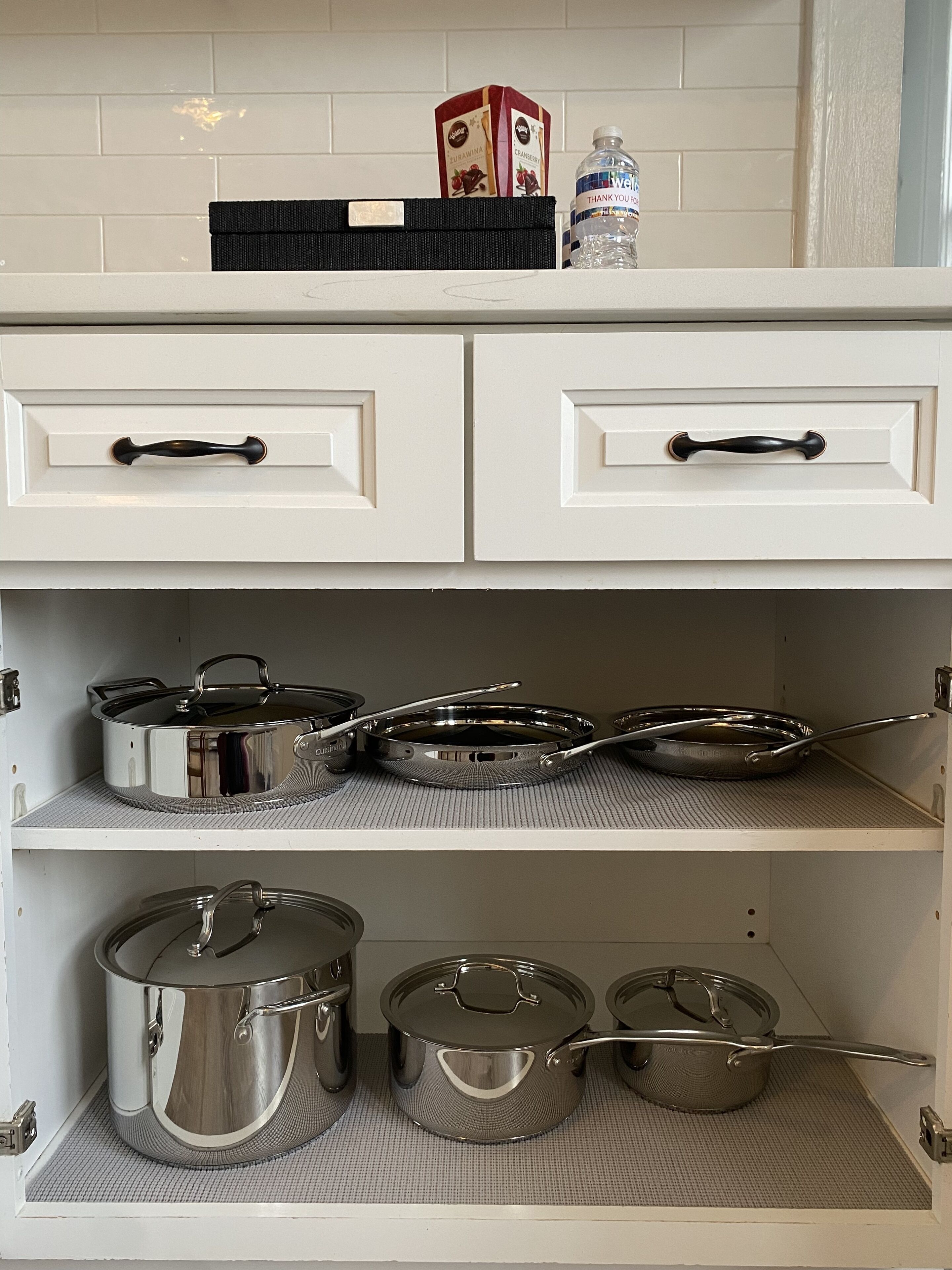 Una cocina con armarios blancos, dos cajones y tres estantes. Se observan dos cajas, una botella de agua y varias ollas y sartenes sobre los estantes.