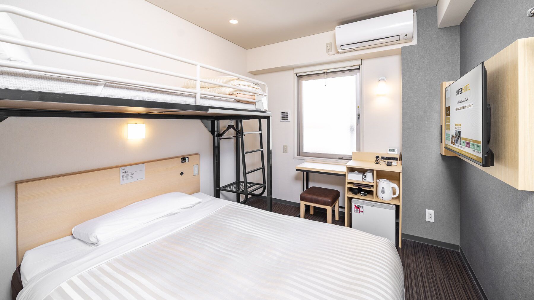 A hotel room with a bunk bed, a desk with a chair, a small table with a kettle, and a wall-mounted TV.