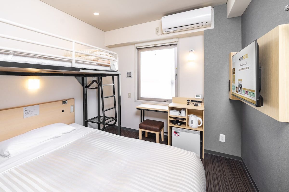Una habitació d'hotel amb un llit superposat, un escriptori amb una cadira, una taula petita amb un bullidor i un televisor fixat a la paret.