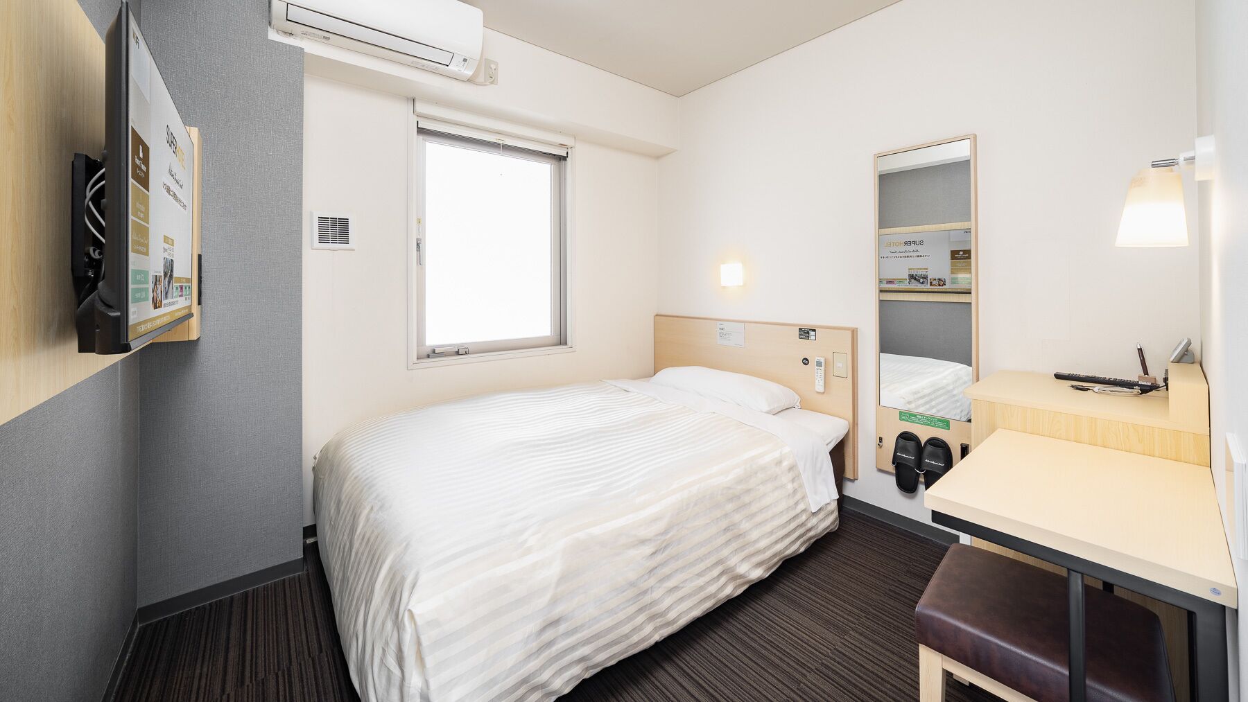 A hotel room with a bed, a desk with a mirror, a shoe rack, and an air conditioning unit.
