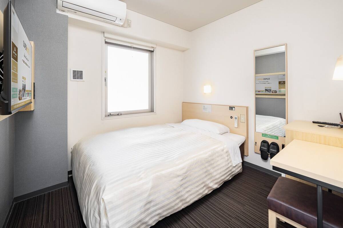 Habitació d'hotel amb un llit, una taula de nit amb mirall, un prestatge per a sabates i un aparell d'aire condicionat.