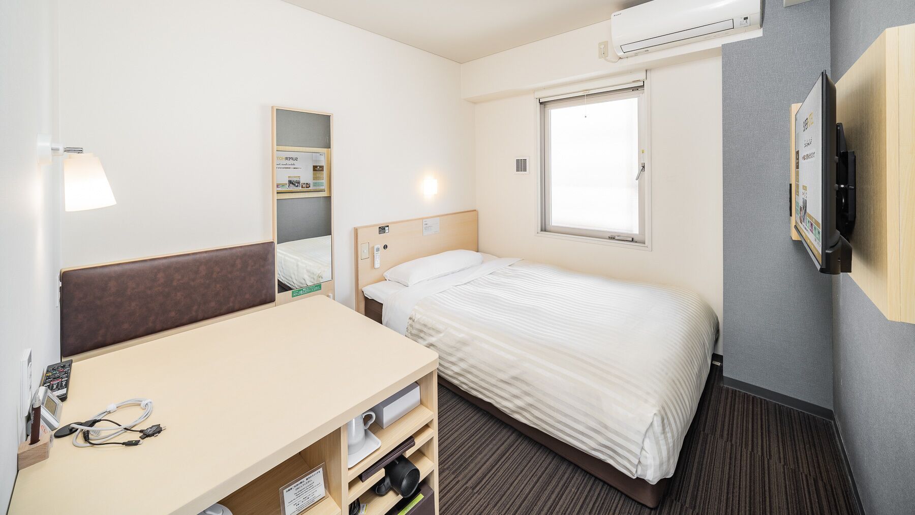 A hotel room with a bed, a desk with a chair, a mirror, a window, and an air conditioning unit.