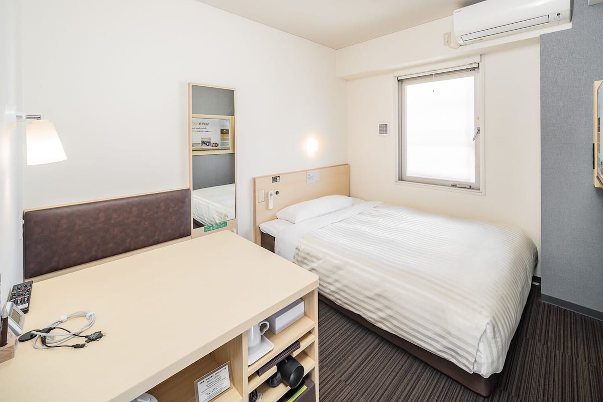 Habitació d'hotel amb un llit, una taula de treball amb cadira, un mirall, una finestra i un aparell d'aire condicionat.