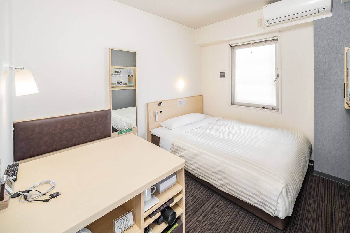 A hotel room with a bed, a desk with a chair, a mirror, a window, and an air conditioning unit.