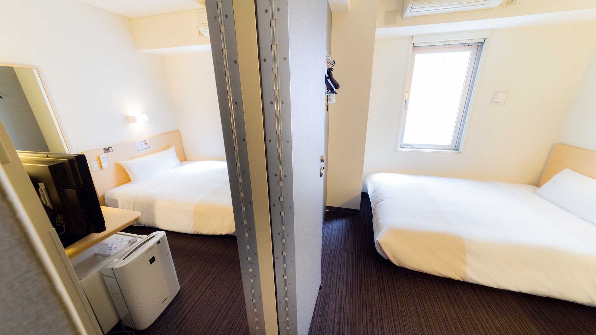 Ein Hotelzimmer mit zwei Betten, einem Schreibtisch mit Computer, einem kleinen Heizgerät und einem Fenster.
