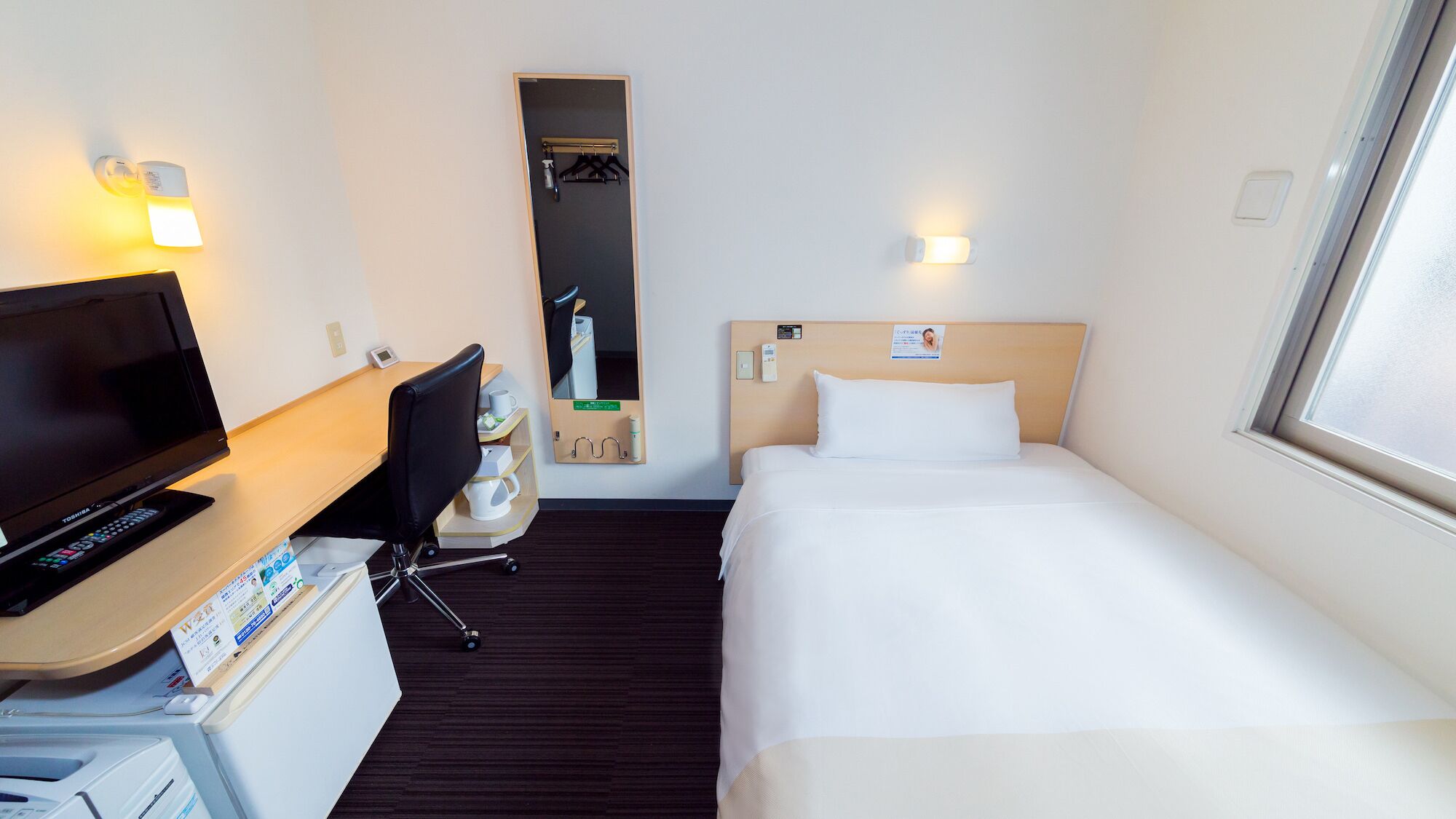 Ein Hotelzimmer mit einem Bett, einem Schreibtisch mit Fernseher, einem Sessel, einem kleinen Tisch, einem Spiegel und einem Fenster.
