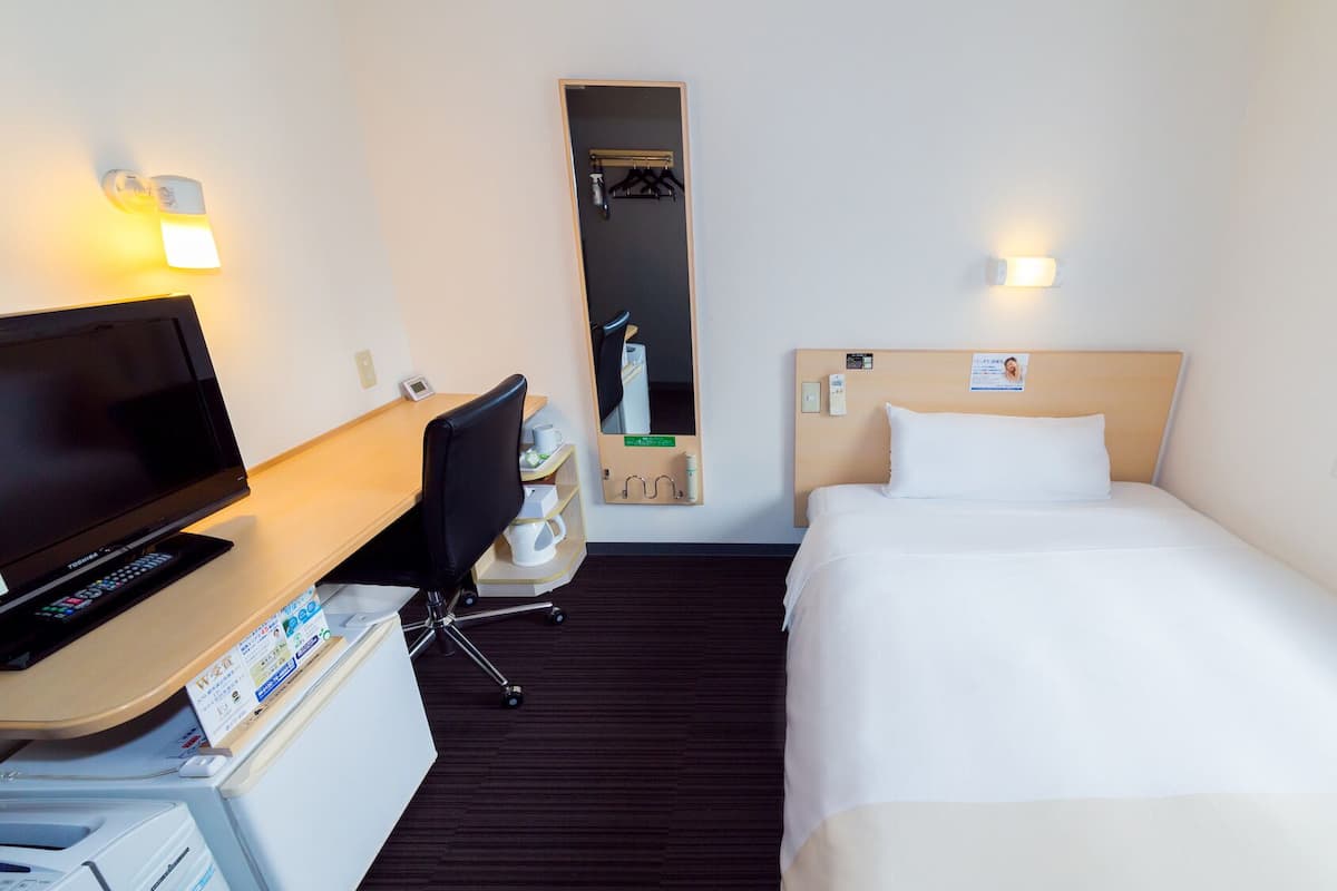 Habitació d'hotel amb un llit, una escriptori amb un televisor, una cadira, una taula petita, un mirall i una finestra.