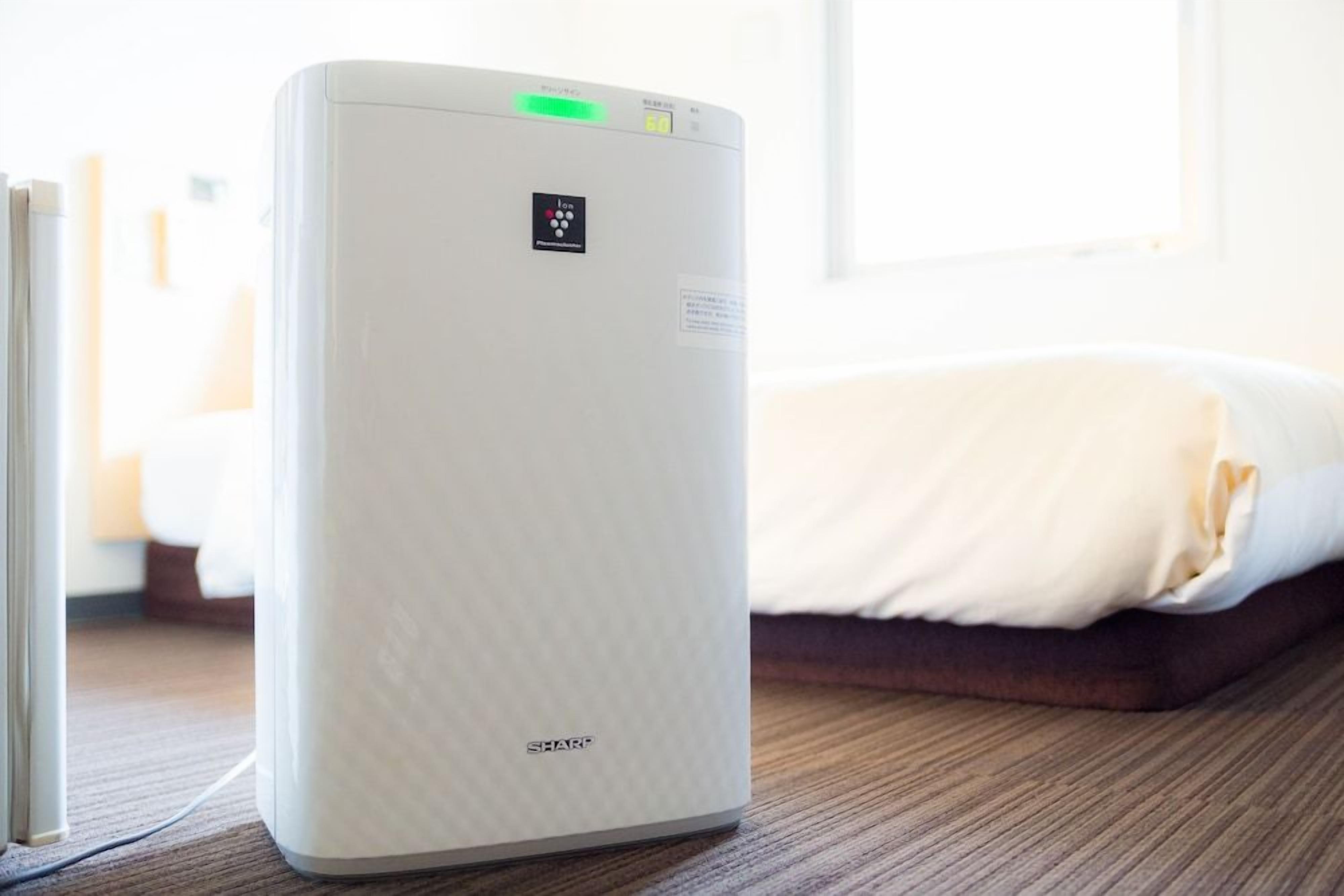 A white portable heater with a control panel, placed on a wooden floor next to a bed with white bedding.