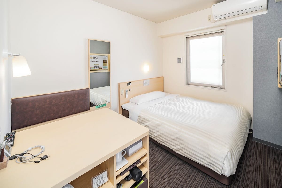 Habitació d'hotel amb un llit, escriptori amb cadira, mirall, finestra i televisió penjada a la paret.