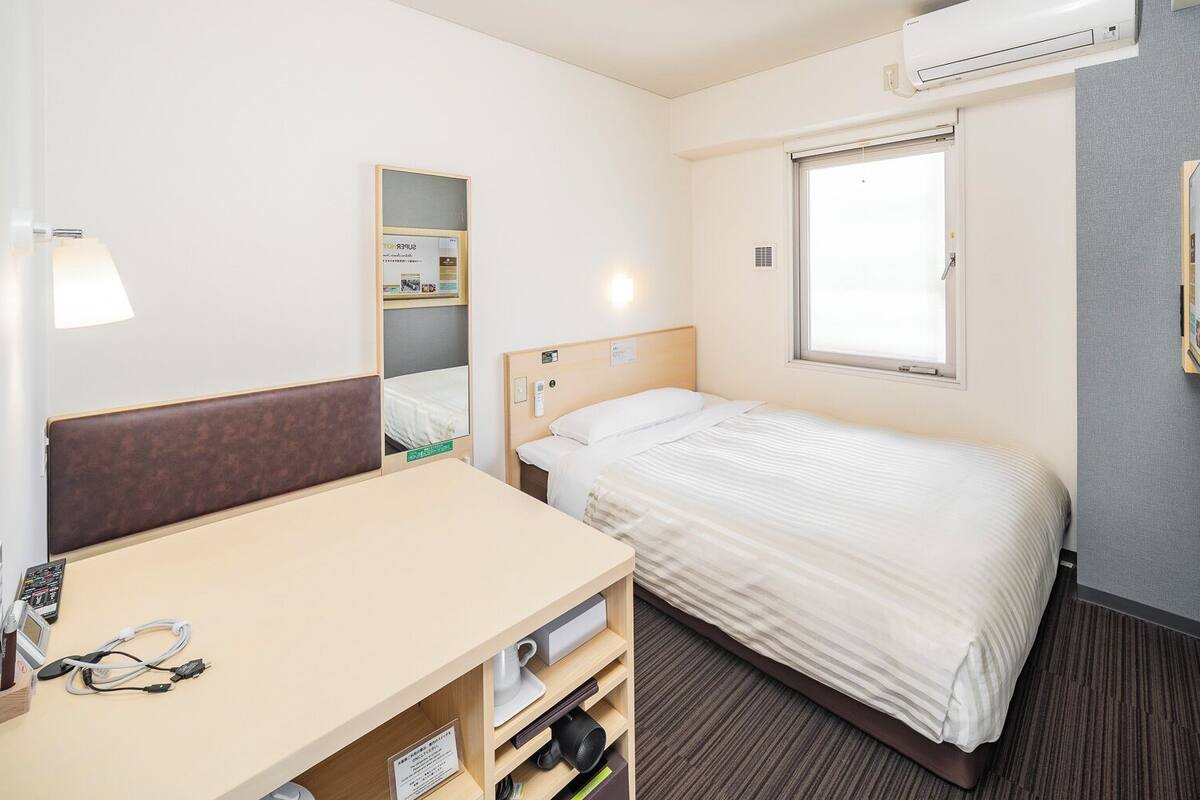 Ein Hotelzimmer mit einem Bett, einem Schreibtisch mit Stuhl, einem Spiegel, einem Fenster und einem an der Wand befestigten Fernseher.