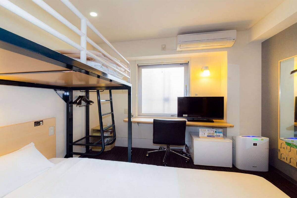 Habitació d'hotel amb un llit superposat, un escriptori amb un ordinador, una televisió i un refrigerador petit.