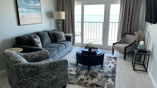 A living room with a sofa, armchair, and coffee table, a TV, and a view of the ocean through a sliding door. - Stunning Views from this lovely 2 bed/2bath (Galveston)