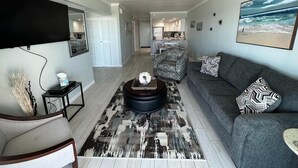 A living room with a grey sofa, a coffee table with a flower arrangement, a patterned rug, and a painting of a beach scene. - Stunning Views from this lovely 2 bed/2bath (Galveston)