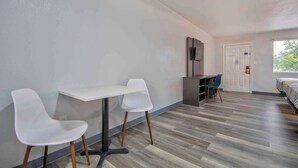 Una habitación con una mesa blanca y dos sillas blancas, un escritorio gris, un televisor en la pared y una cama con ropa de cama a rayas.