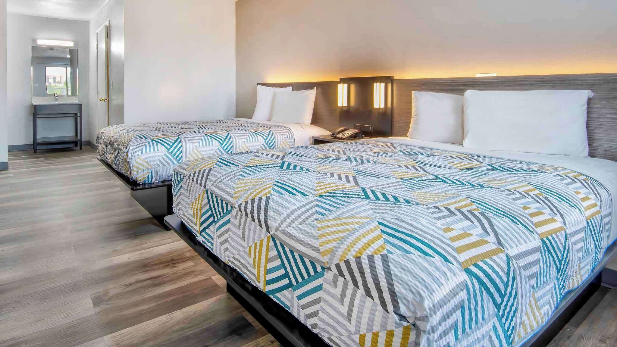 Habitación de hotel con dos camas, una mesita de noche con lámpara, y un baño que se ve al fondo.