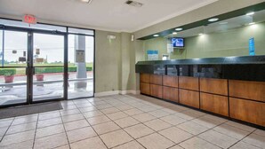 Un área de recepción con entrada por puerta de vidrio, un mostrador de recepción con pantalla digital y piso de baldosas.
