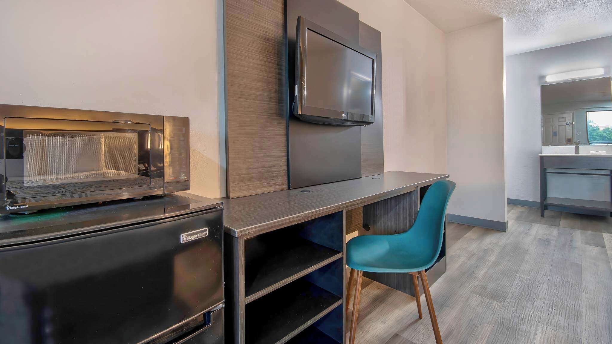 Eine kompakte Küchenzeile mit Mikrowelle, kleinem Kühlschrank, einem flachbildfernseher an der Wand und einem modernen blauen Stuhl.