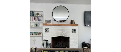 A fireplace with a white brick surround, a mirror above it, and a shelf with books and decorative items.