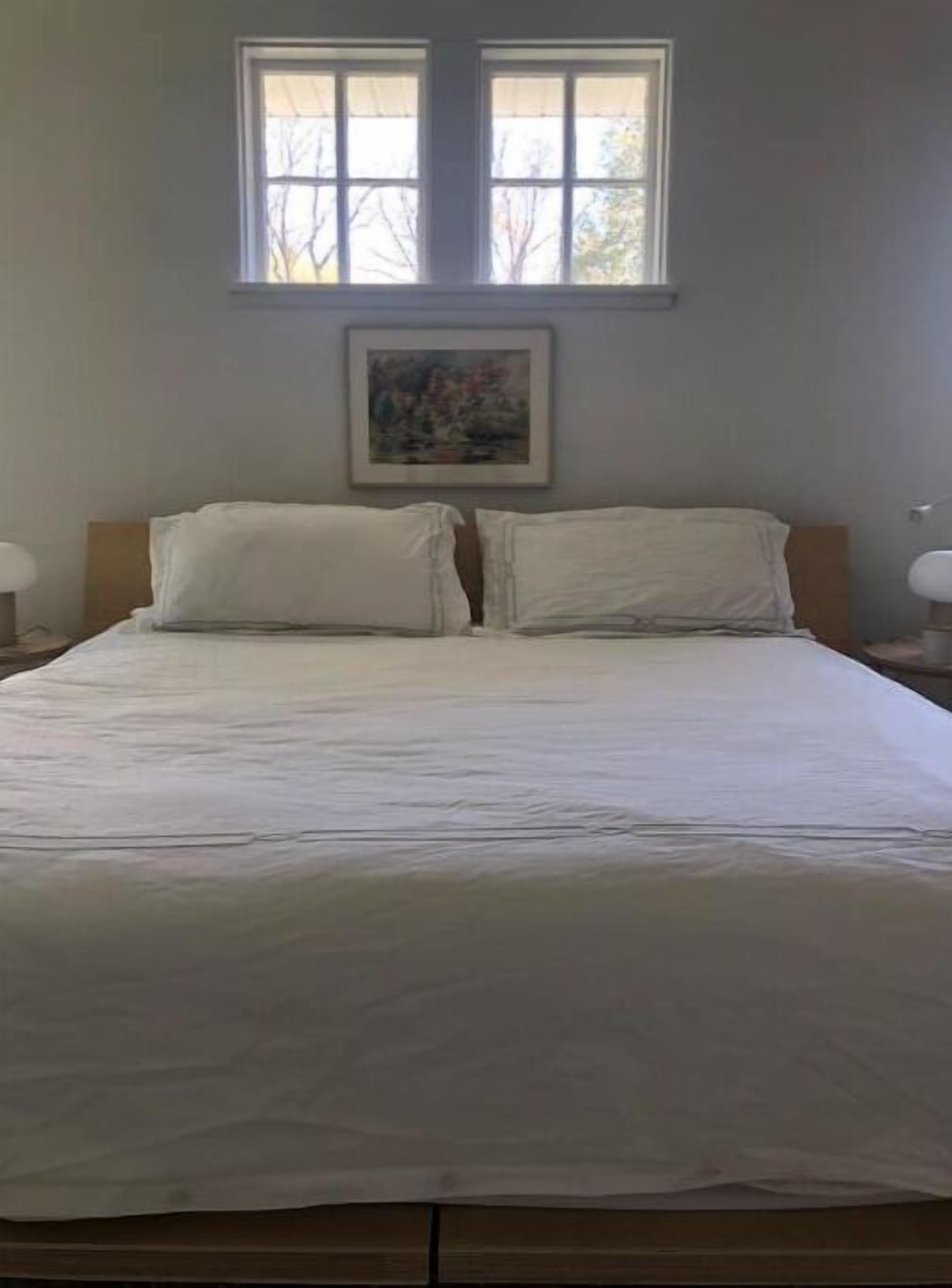 Un lit bien fait, avec du linge de lit blanc, dans une pièce dotée d’une grande fenêtre et d’un tableau encadré au mur.