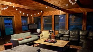 Un salon chaleureux avec un canapé, des fauteuils et une table basse. De grandes fenêtres et un plafond en bois complètent l’ensemble.