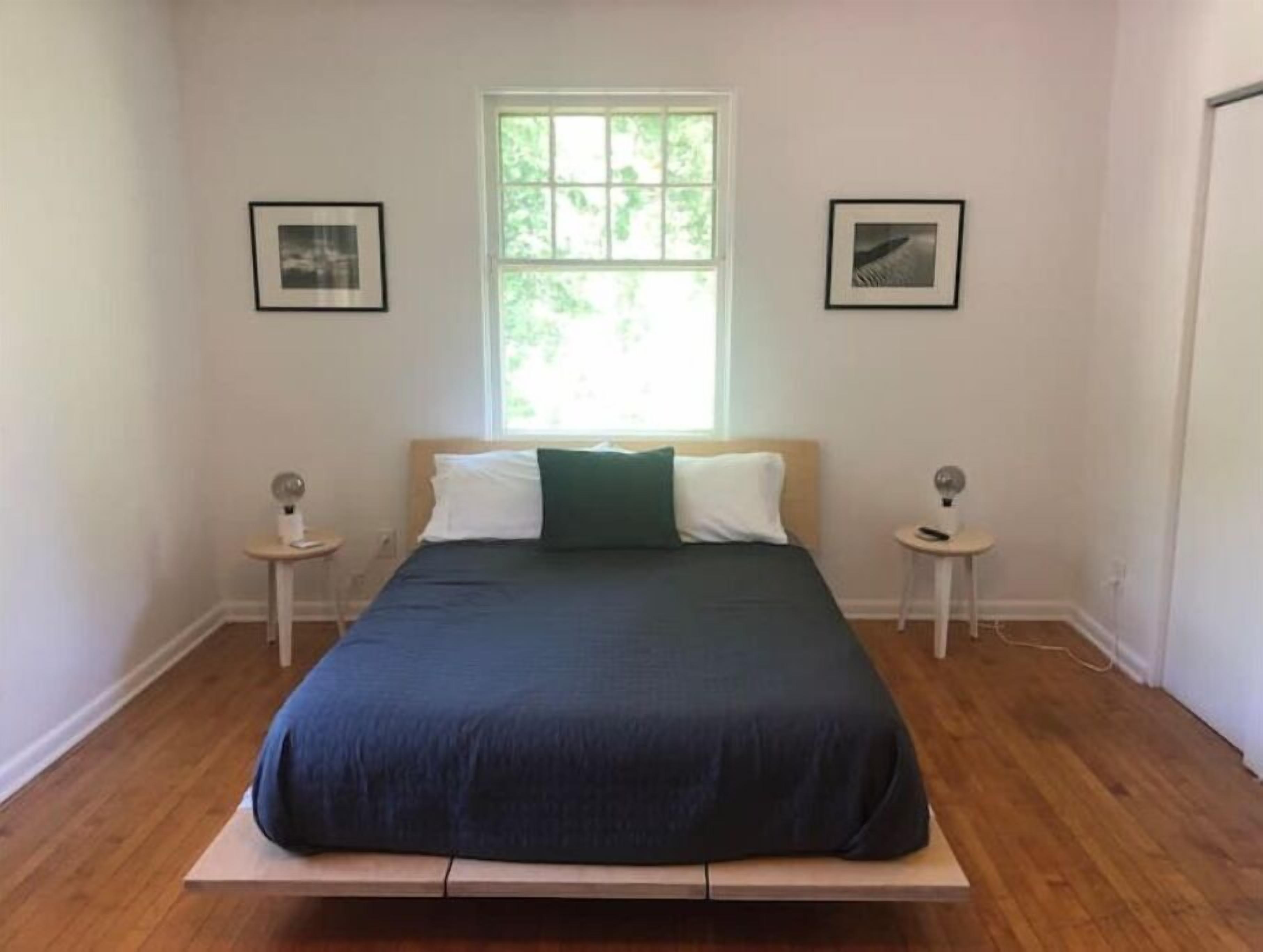 A bedroom with a bed centered under a window, flanked by two small bedside tables with lamps, and framed pictures on the walls.