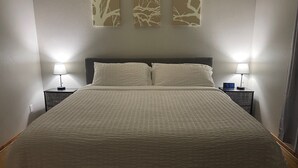 Una cama bien hecha con un edredón blanco texturizado, dos lámparas de noche y un reloj digital en la mesita.
