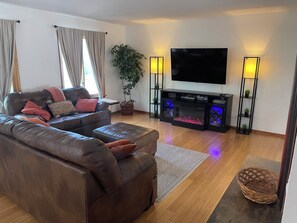 A living room with a leather sofa, a flat-screen TV, a fireplace with blue lighting, and a potted plant.