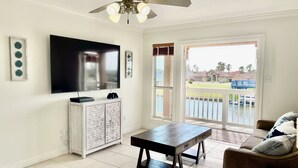 A living room with a flat-screen TV, a ceiling fan, a wooden cabinet, a coffee table, and a view of a water body and houses through a glass door. - Spectacular Sunsets! Private Pool & Spa on Canal - Walk to Beach & Restaurants!! (Corpus Christi)