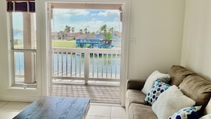 A room with a view of a lake, a balcony with a blue deck, and a beige sofa with patterned pillows. - Spectacular Sunsets! Private Pool & Spa on Canal - Walk to Beach & Restaurants!! (Corpus Christi)