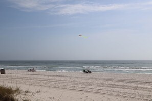 Ein Strand mit blauem Himmel, einigen entspannten Personen und einem fliegenden Drachen.