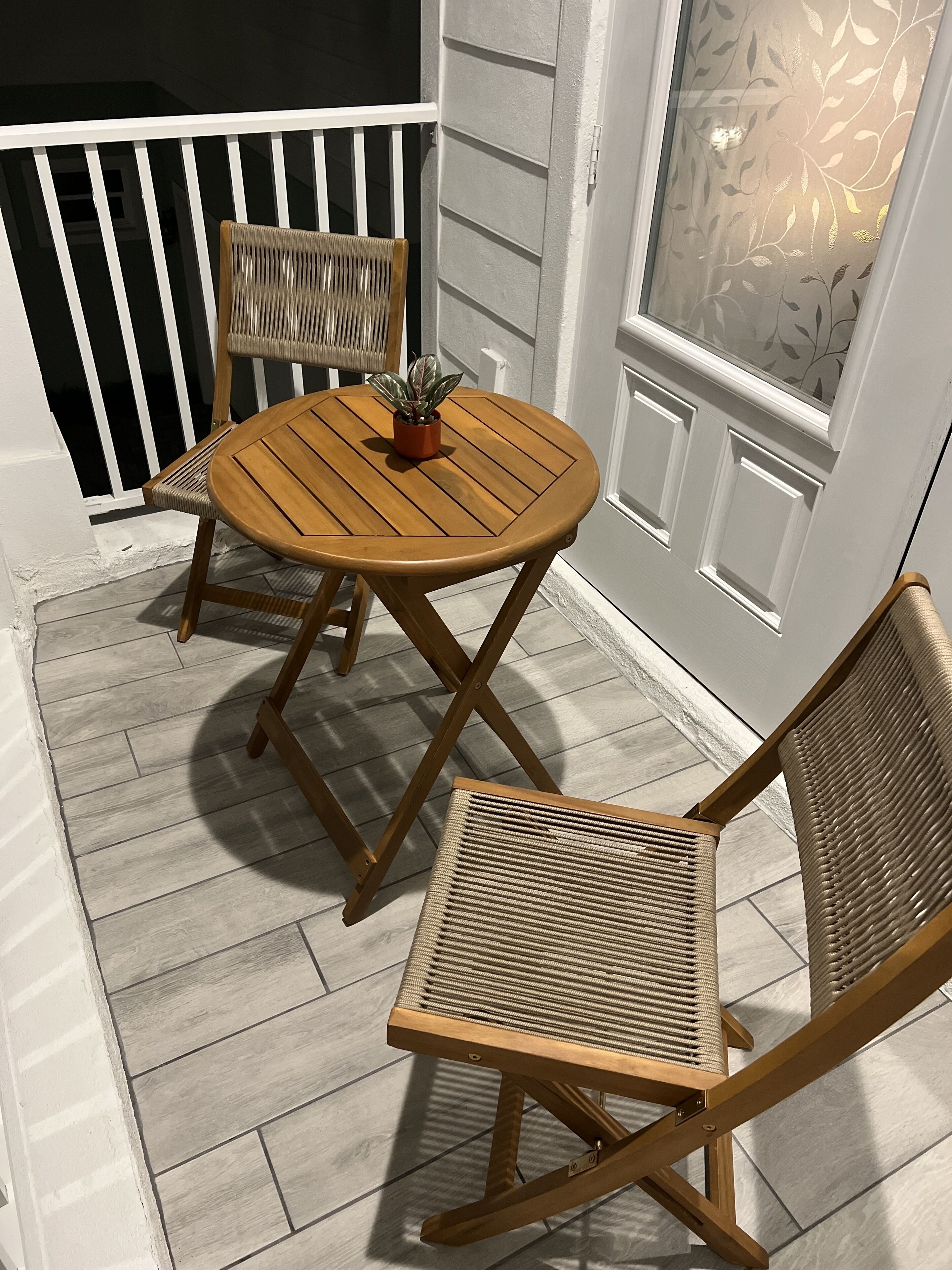 Un espace extérieur aménagé avec des meubles en bois, comprenant une table ronde et deux chaises, un pot de fleurs et une porte blanche dotée d’une fenêtre en verre dépoli.