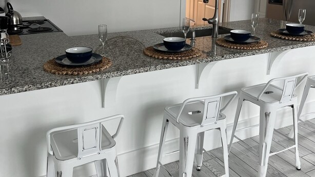 Una cocina moderna con armarios blancos, encimera de granito y una barra integrada con taburetes blancos.