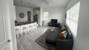 Una sala de estar moderna con un sofá gris, un televisor y una cocina con taburetes blancos.