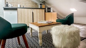 Ett mysigt sovrum på vinden med ett sluttande tak, ett träbord, en grön stol, en fluffig vit hund och en liten köksdel med mikrovågsugn och kylskåp.
