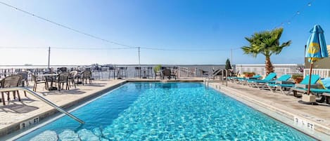 A rectangular pool with clear blue water, surrounded by lounge chairs and tables, with a clear sky above.