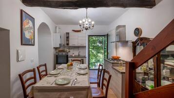 Un comedor con una mesa preparada para cuatro personas, una araña y vistas a la cocina y al follaje exterior.