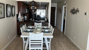 A dining area with a wooden table set for four, surrounded by white chairs. A chandelier hangs above, and the room has a neutral color scheme with beige walls and a wooden floor.