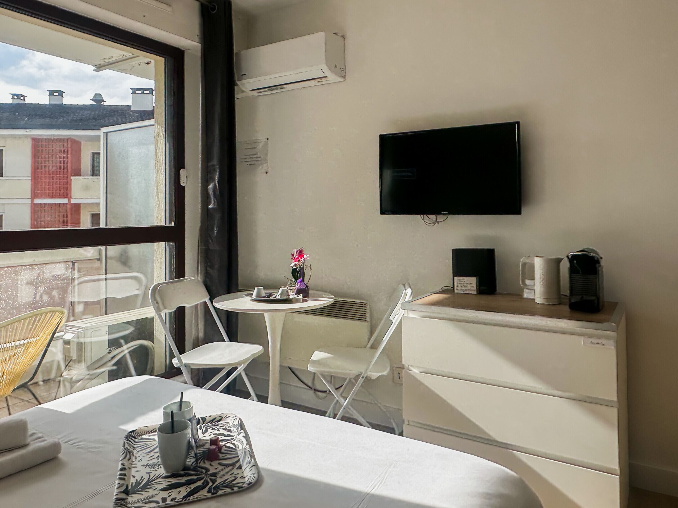 Una habitación de hotel con cama, una mesita con un jarrón de flores, un televisor montado en la pared y una cafetera sobre una encimera.