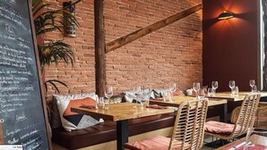 Ein Restaurant mit Backsteinwänden, Holzbalken und Korbstühlen. Die Tische sind mit Weingläsern und Speisekarten gedeckt.