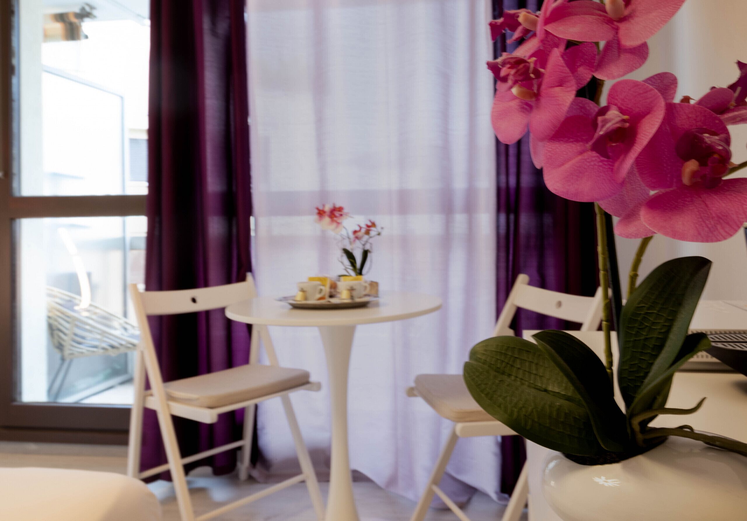 Une pièce avec une table ronde, deux chaises blanches, un vase contenant des orchidées roses et une fenêtre ornée de rideaux violets.