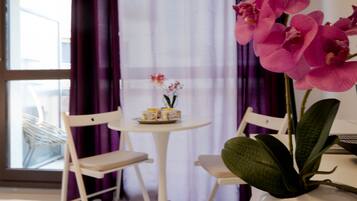 Une pièce avec une table ronde, deux chaises blanches, un vase contenant des orchidées roses et une fenêtre ornée de rideaux violets.