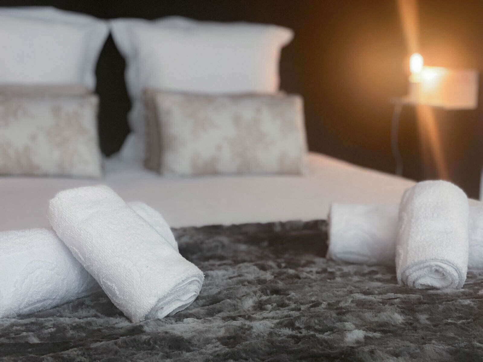 Cama bem arrumada com lençóis e almofadas brancas, tapete escuro com textura e abajur com luz quente.
