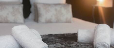 Cama bem arrumada com lençóis e almofadas brancas, tapete escuro com textura e abajur com luz quente.
